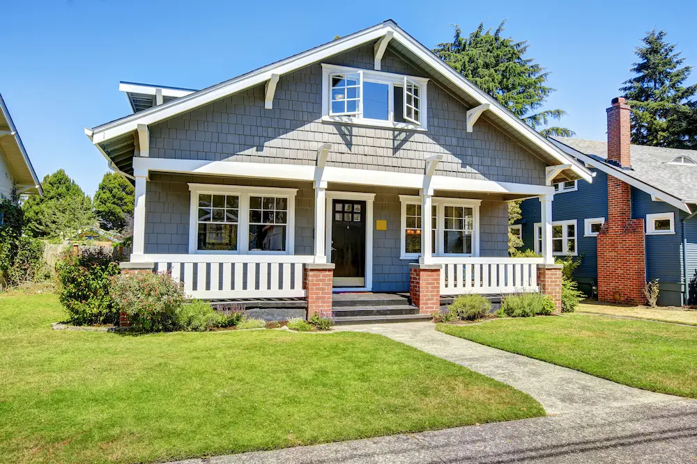 Gray Craftsman house with a brick chimney, white trim, and a covered porch. The manicured lawn and sunny setting create a welcoming feel.
